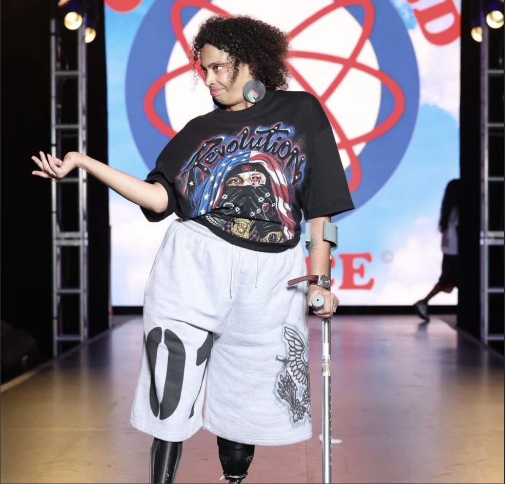 Eman Rimawi-Doster on a fashion runway, smiling over her shoulder, wearing a Revolution T-shirt and shorts with a forearm crutch, her two prosthetic legs visible, in front of a One World Peace backdrop.